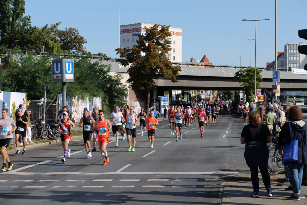 Gruppe von Menschen, die bei einem Marathon auf einer von Bäumen, Laternen, Schildern, Fahrrädern, einem Zaun, Gras, einer Brücke, Gebäuden mit Fenstern und einem klaren blauen Himmel gesäumten Straße laufen.