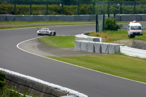 Ein Rennwagen fährt auf einer Rennstrecke mit einem Polizeiwagen im Hintergrund, umgeben von Gras, Pflanzen, Bäumen und einem Zaun, mit einem Fahrzeug auf der rechten Seite und Text unten.