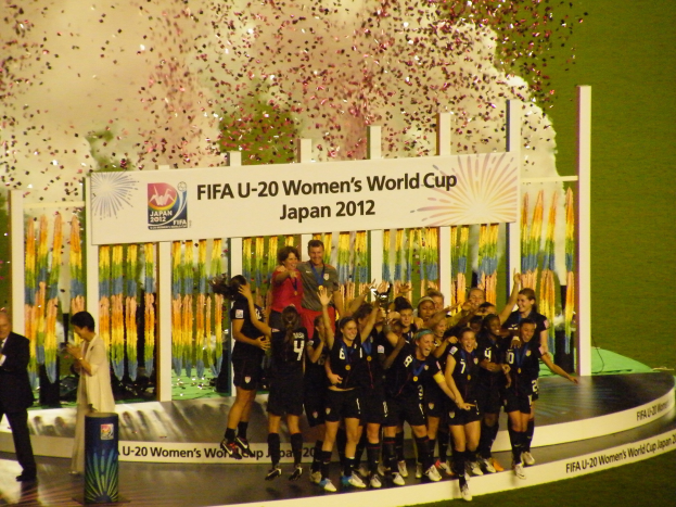 Gruppe von Frauenfußballspielerinnen in schwarzer Uniform auf einer Bühne feiernd umgeben von einer Menge, mit einer Tafel und dekorativen Elementen im Hintergrund.