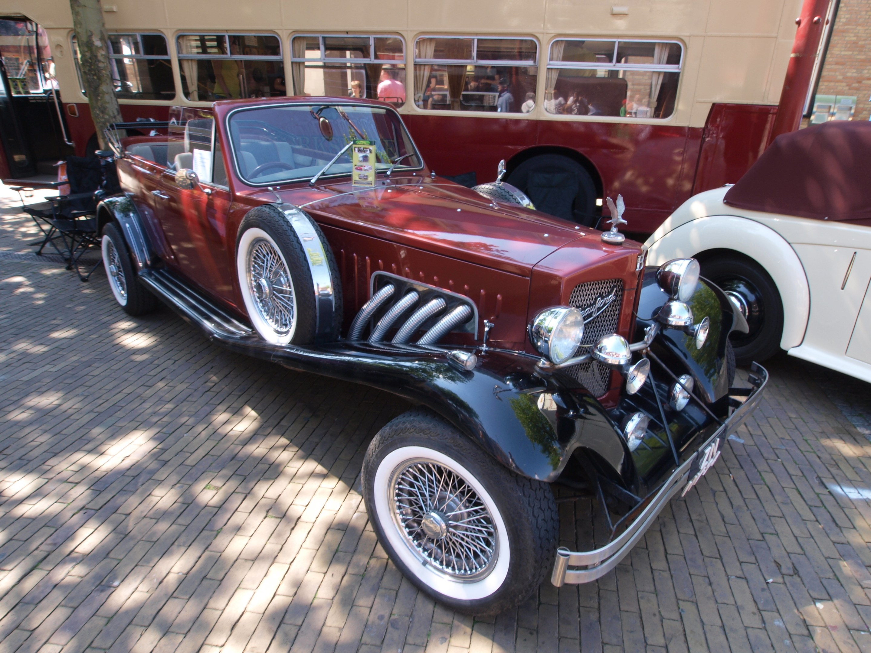 Vintage-Auto neben einem Doppeldeckerbus auf einem Kopfsteinpflasterweg geparkt; Stühle und ein Baum im Vordergrund und ein Gebäude im Hintergrund; Personen im Bus sichtbar.