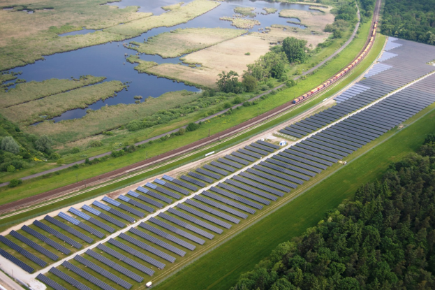 Luftaufnahme einer Solar-Farm mit Panels auf einem Feld, umgeben von Bäumen, Gras, Pflanzen und Wasser, mit einem Zug auf einer nahen Bahnstrecke.