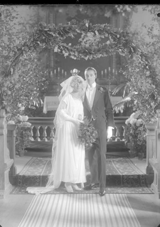 Schwarz-weißes Foto eines Mannes und einer Frau vor einem Blumenschmuckbogen, die Frau hält einen Strauß.