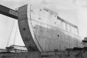 Schwarzes und weißes Foto einer großen Betonstruktur im Bau, mit einer Wand unten, einer Brücke links und einem Himmel im Hintergrund.