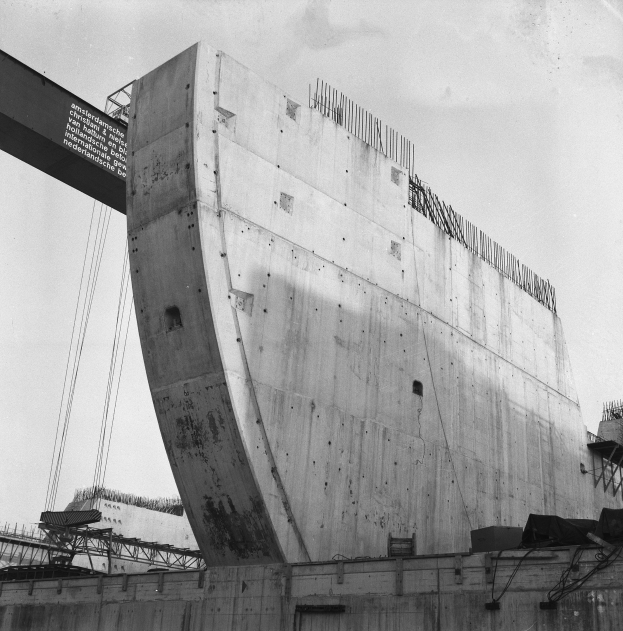 Schwarzes und weißes Foto einer großen Betonstruktur im Bau, mit einer Wand unten, einer Brücke links und einem Himmel im Hintergrund.