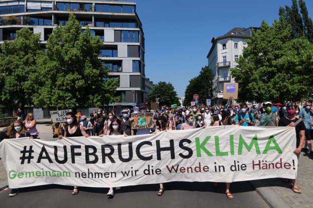 Gruppe von Menschen mit Masken, die ein Transparent mit der Aufschrift 'Aufbruchsklima' vor einem Gebäude mit Bäumen und klarem Himmel halten.