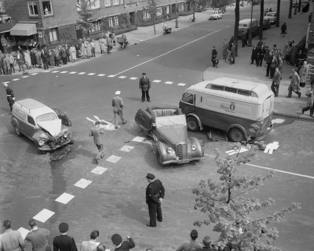 Schwarz-weißes Bild einer Autounfall-Szene an einer Straßenkreuzung mit einem Lieferwagen, anderen Fahrzeugen und einer Gruppe von Menschen, mit Gebäuden, Bäumen, Pfählen und Texttafeln im Hintergrund.