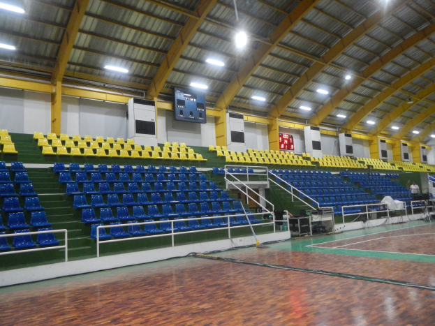 Indoor-Sporthalle mit Holzboden, blauen und gelben Sitzplätzen, Geländern, Treppen zum Sitzbereich, wandmontierten Bildschirmen, Deckenleuchten und einigen Menschen.