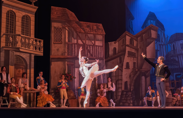 Zwei Tänzer performen auf einer Bühne im Royal Opera House in London, mit einem Publikum im Hintergrund und einem Bildschirm, der Gebäudeabbildungen anzeigt.