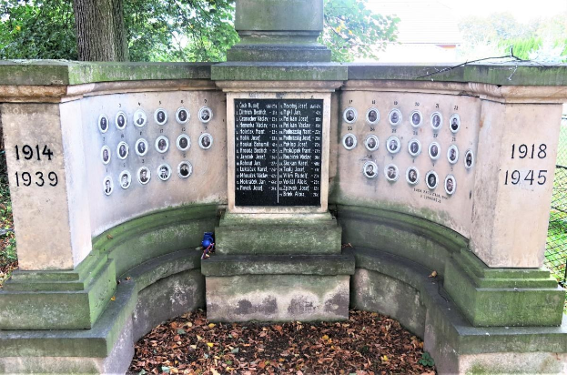Ein Holocaust-Gedenkdenkmal in einem jüdischen Friedhof in Berlin, das eine Wand mit Text und Zahlen zeigt, umgeben von Bäumen, einem Zaun und verstreuten trockenen Blättern.