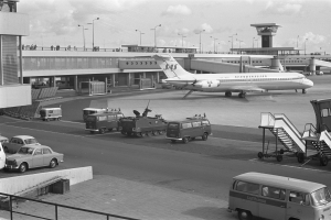 Ein Schwarz-Weiß-Foto eines Flughafens mit Autos und Lastwagen auf der Straße, einem Flugzeug auf dem Bahnsteig, Treppen mit Geländern, einem Gebäude mit Säulen und Geländern im Hintergrund, Laternenmasten und einem bewölkten Himmel.