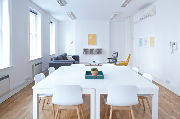 Innenraum eines Apartments in Berlin mit einem weißen Tisch mit einer Pflanze in einer Schale, Stühlen, einer Couch mit Kissen, einer Lampe, Regalen mit Gegenständen, einer Klimaanlage, Fotorahmen an der Wand, Fenstern und Deckenleuchten.