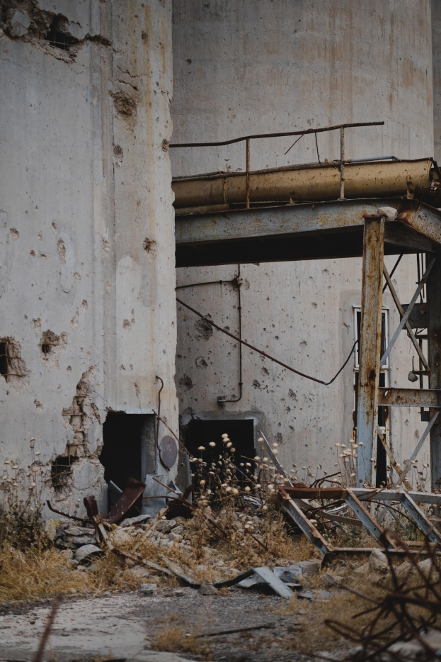 Altes Gebäude mit bröckelnden Wänden, zerbrochenen Fenstern und einem großen zentralen Metallgerüst, umgeben von Pflanzen, Eisenstangen und Schutt.