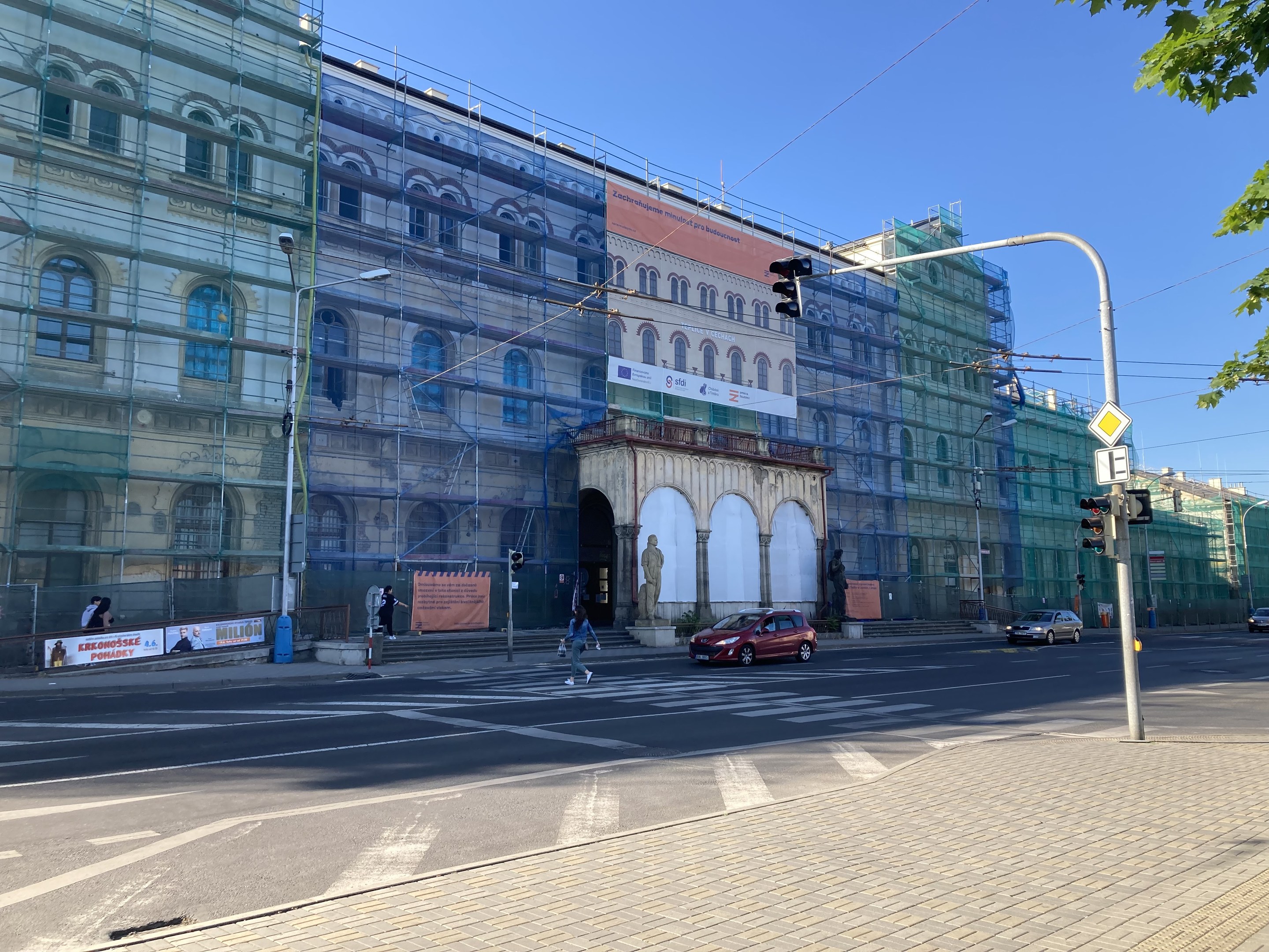 Großes Gebäude mit Gerüst während der Renovierung, umgeben von Fahrzeugen, Passanten, Ampeln, Schildern, Straßeninfrastruktur, Bäumen und einem klaren blauen Himmel.