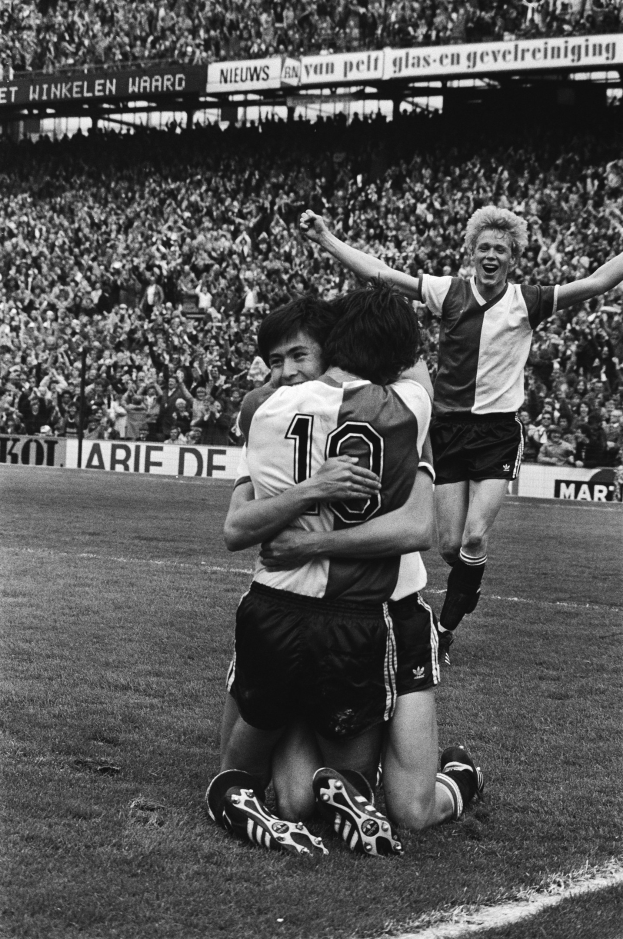 Drei Fußballspieler, die sich nach einem Tor auf dem Boden umarmen, vor einem jubelnden Publikum und schwenkenden Fahnen im Hintergrund, in einer schwarz-weißen Stadionatmosphäre.