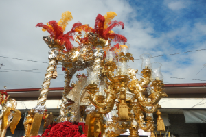 Ein großer goldener und roter Festwagen mit Blumen und anderen Schmuckelementen bei einem Karnevalsumzug, mit einem Gebäude, Strommasten mit Drähten und einem bewölkten Himmel im Hintergrund.