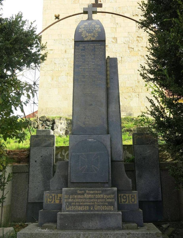 Denkmal für deutsche Soldaten, die im Zweiten Weltkrieg starben, vor einer Kirche mit einem Kreuz, umgeben von Bäumen und einer Mauer.