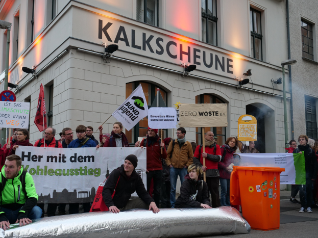Gruppe von Menschen bei einer Demonstration vor einem Gebäude in Deutschland, die Schilder und Plakate halten, mit zwei Personen im Vordergrund und einem Müllcontainer auf der rechten Seite.