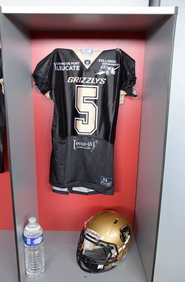 Ein Fußballtrikot und Helm hängen an einer Spindtür, mit einer Wasserflasche auf dem Regal darunter; das Trikot trägt das Logo der Mannschaft.