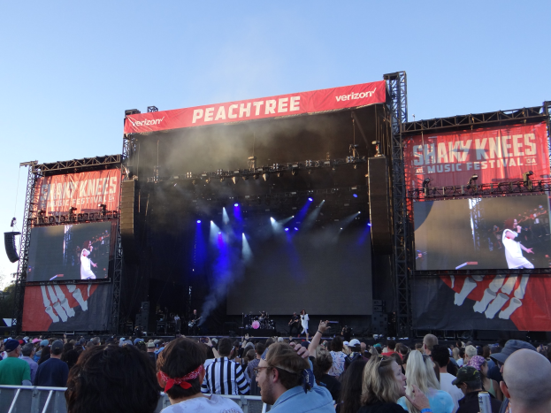 Eine große Menge steht vor einer hell erleuchteten Bühne auf dem Shaky Knees Music Festival, wo Musiker hinter Bannern spielen, mit einem Geländer im Vordergrund und einem klaren blauen Himmel im Hintergrund.