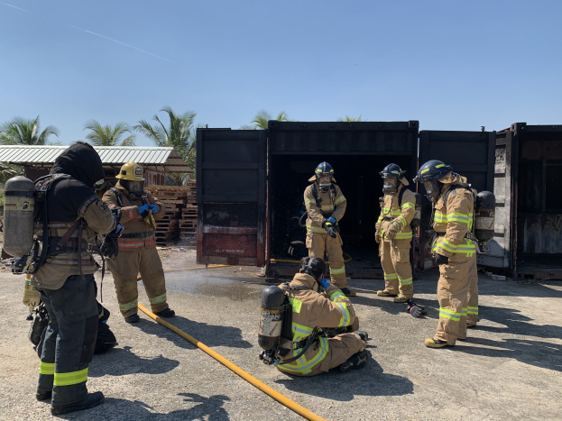 Feuerwehrleute in Helmen und Ausrüstung vor einem Gebäude mit Rohren stehend, mit Bäumen, Schuppen und hölzernen Gegenständen im Hintergrund bei einer scheinbar Feuerlöschtraining.