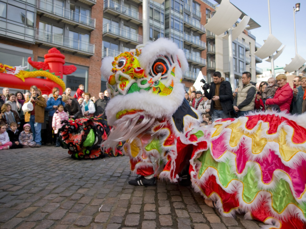 Ein lebhaftes chinesisches Neujahrsfest in Amsterdam mit einer Drachenvorführung vor einer Zuschauermenge, einige machen Fotos, vor einer Kulisse aus Gebäuden, Laternenmästen und einem klaren blauen Himmel.