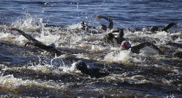 Gruppe von Menschen, die synchron beim Triathlon schwimmen, tragen schwarze Neoprenanzüge und Helme, mit Sonnenreflexion auf dem Wasser.