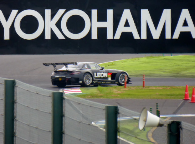 Ein schwarzer Sportwagen fährt auf einer Rennstrecke mit einem "Yokohama"-Schild im Hintergrund, umgeben von Verkehrskegeln, Gras und einem Zaun, mit einem Lautsprecher im Vordergrund.