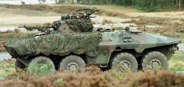 Militärischer Mannschaftstransporter mit montierter großer Waffe, der auf einer Straße neben einem Wald fährt.