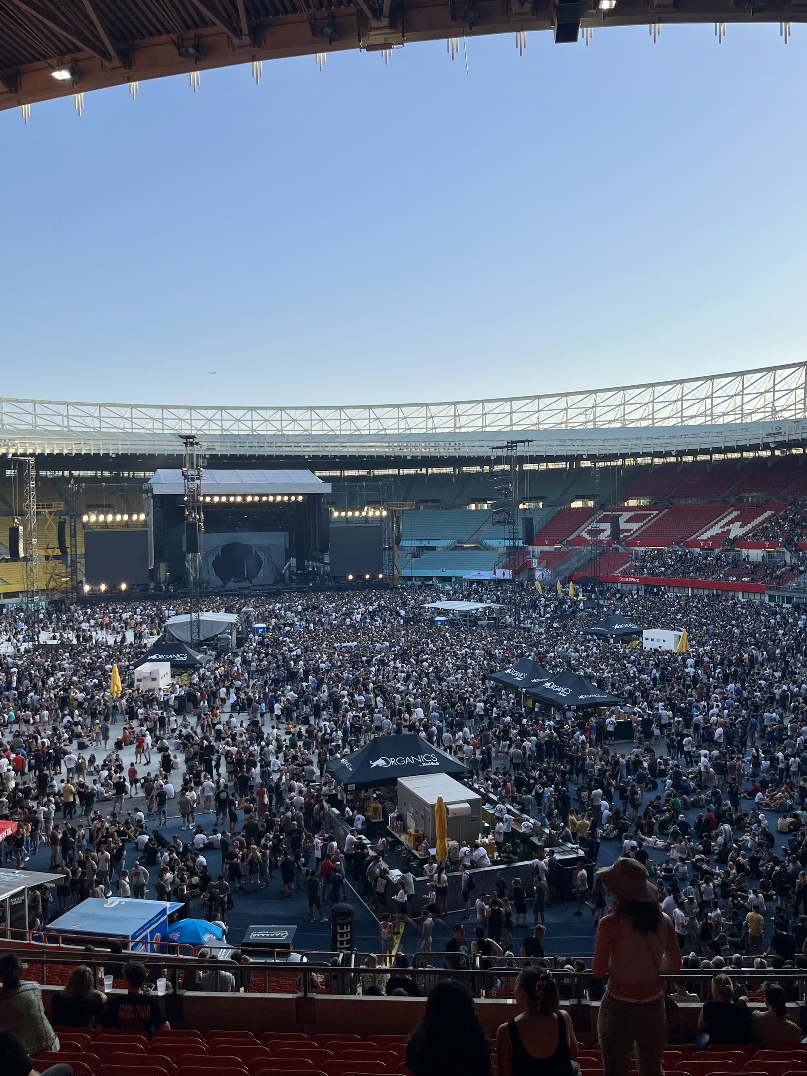Eine große Menschenmenge sitzt und steht in einem stadionähnlichen Veranstaltungsort, umgeben von Geländern, Zelten und Beleuchtungsausrüstung, mit einem Dach und Lichtern oben und dem Himmel im Hintergrund.