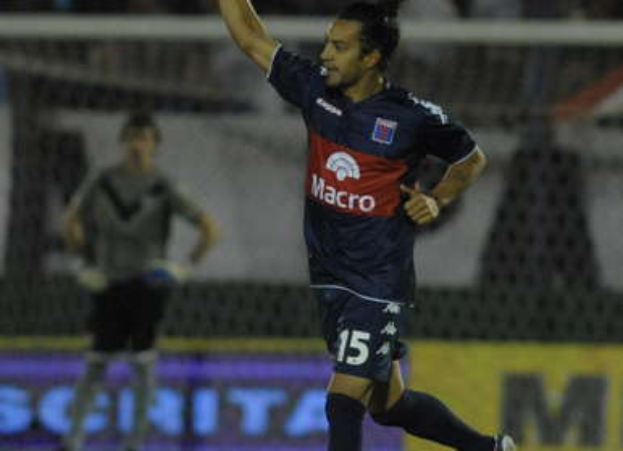 Ein Fußballspieler feiert auf dem Boden nach einem Tor, mit einer jubelnden Menge und einer Person im Hintergrund und einer Tafel mit Text dahinter, alles vor einem unscharfen, energischen Hintergrund.