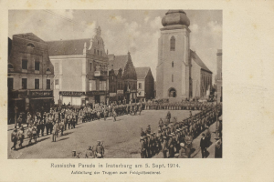Ein Schwarz-Weiß-Foto einer Parade in Insterburg am 5. September 1914 mit vielen Menschen, Gebäuden und einem bewölkten Himmel sowie einem Text am unteren Rand.