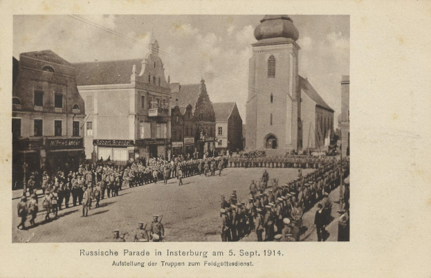 Ein Schwarz-Weiß-Foto einer Parade in Insterburg am 5. September 1914 mit vielen Menschen, Gebäuden und einem bewölkten Himmel sowie einem Text am unteren Rand.
