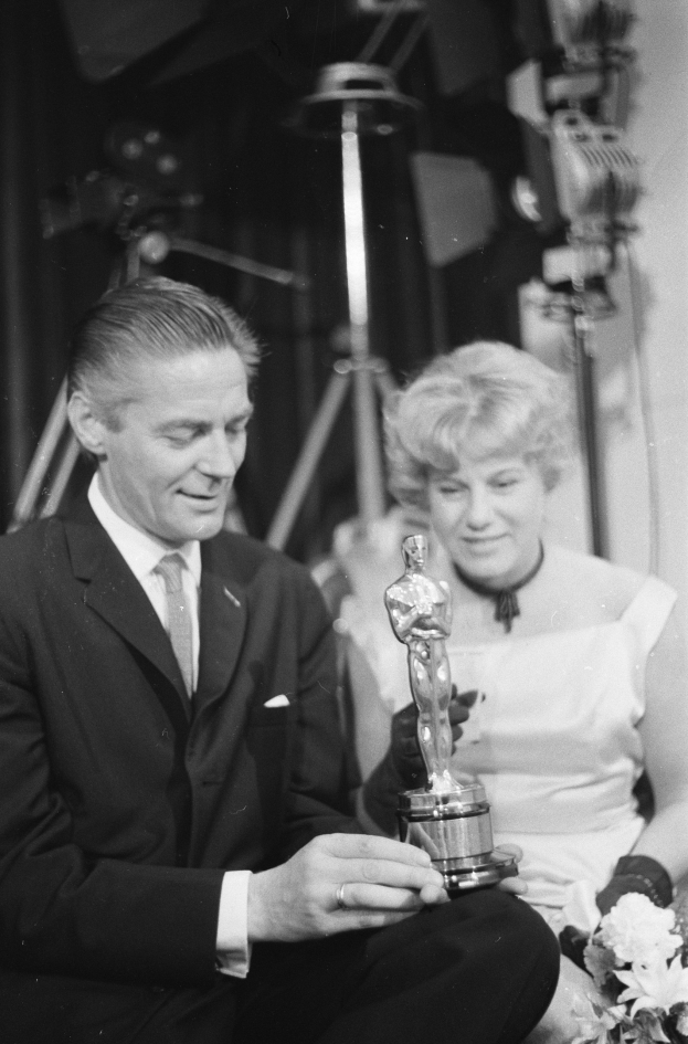 Ein Mann in einem schwarzen Blazer, weißem Hemd und Krawatte hält eine Oscar-Statue in der Hand, während er neben einer Frau in Abendgarderobe sitzt, die einen Blumenstrauß hält, mit Hintergrundbeleuchtung in einem Schwarz-Weiß-Bild.
