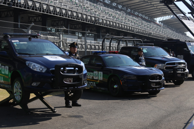Eine Gruppe von Polizeiwagen vor einer Rennstrecke geparkt, mit uniformierten Beamten auf der Straße und einem Stadion mit Geländern und Maschendrahtzaun im Hintergrund.