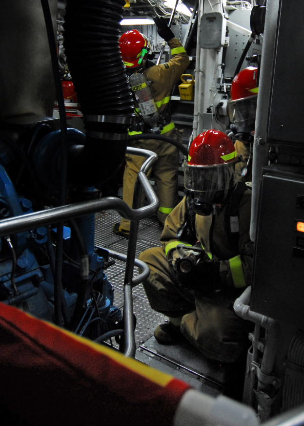 Feuerwehrleute in Helmen und Schutzausrüstung arbeiten an einem Löschwagen mit Rohren und Gegenständen im Hintergrund, unter einem Licht oben.