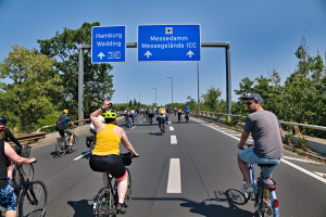 Eine Gruppe von Radfahrern in Helmen fährt auf einer Straße mit einer Begrenzung auf einer Seite und Bäumen auf der anderen unter einem klaren blauen Himmel mit Laternen im Hintergrund; ein Schild oben zeigt eine Radtour in Hamburg.