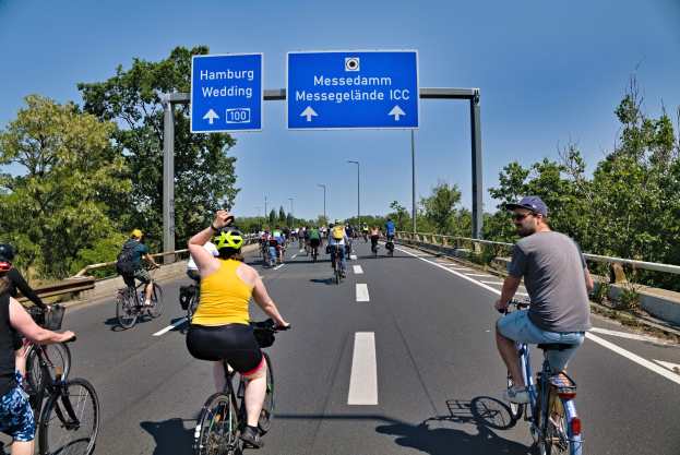 Eine Gruppe von Radfahrern in Helmen fährt auf einer Straße mit einer Begrenzung auf einer Seite und Bäumen auf der anderen unter einem klaren blauen Himmel mit Laternen im Hintergrund; ein Schild oben zeigt eine Radtour in Hamburg.
