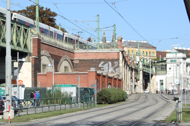 Stadtstraßenszene mit einem Zug auf Schienen, umgeben von Gebäuden, Straßeninfrastruktur, Fahrzeugen, Füßgängern, Vegetation und einem klaren Himmel.