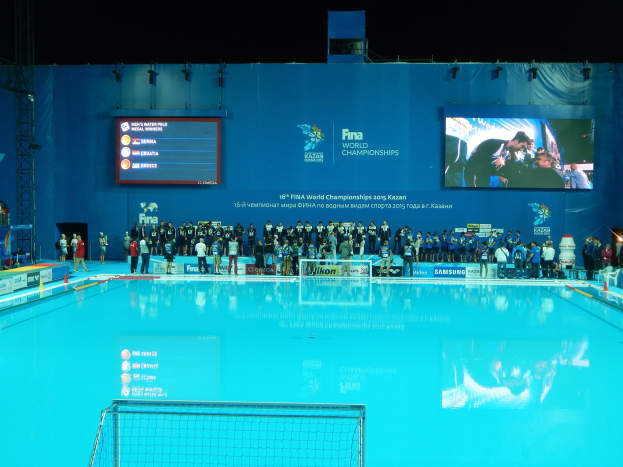 Gruppe von Menschen, die um ein Hallenbad mit Netz am Boden stehen, mit einem Bildschirm im Hintergrund, der "FINA World Championships 2015 Kazan" anzeigt.