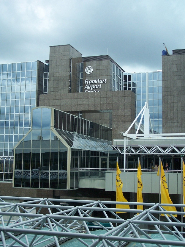 Frankfurt Airport Gebäude mit Glaswänden, Text, gelben Flaggen und Eisenstangen vor einem bewölkten Himmel.
