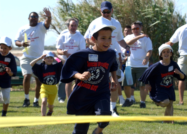 Kinder bei einem Lauf auf einer grünen Wiese mit einem gelben Band im Vordergrund, einige tragen Mützen und Schutzbrillen, andere halten Gegenstände, vor Bäumen und einem klaren blauen Himmel.