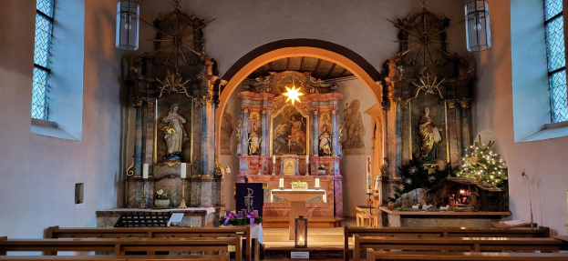 Innenraum einer Kirche mit hölzernen Kirchenbänken, Kerzenständern, Blumenbouquets, Statuen, Lichtern, Fenstern und einem geschmückten Weihnachtsbaum im Hintergrund unter einem Bogen.