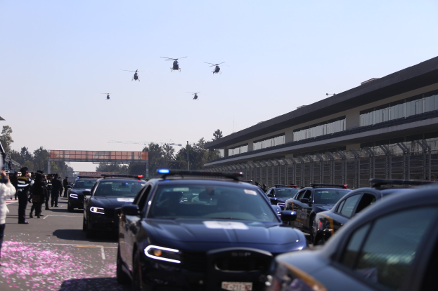 Polizeifahrzeuge vor einem Gebäude geparkt, Menschen auf der linken Seite, Hubschrauber in der Luft und eine Brücke im Hintergrund bei klarem blauem Himmel.