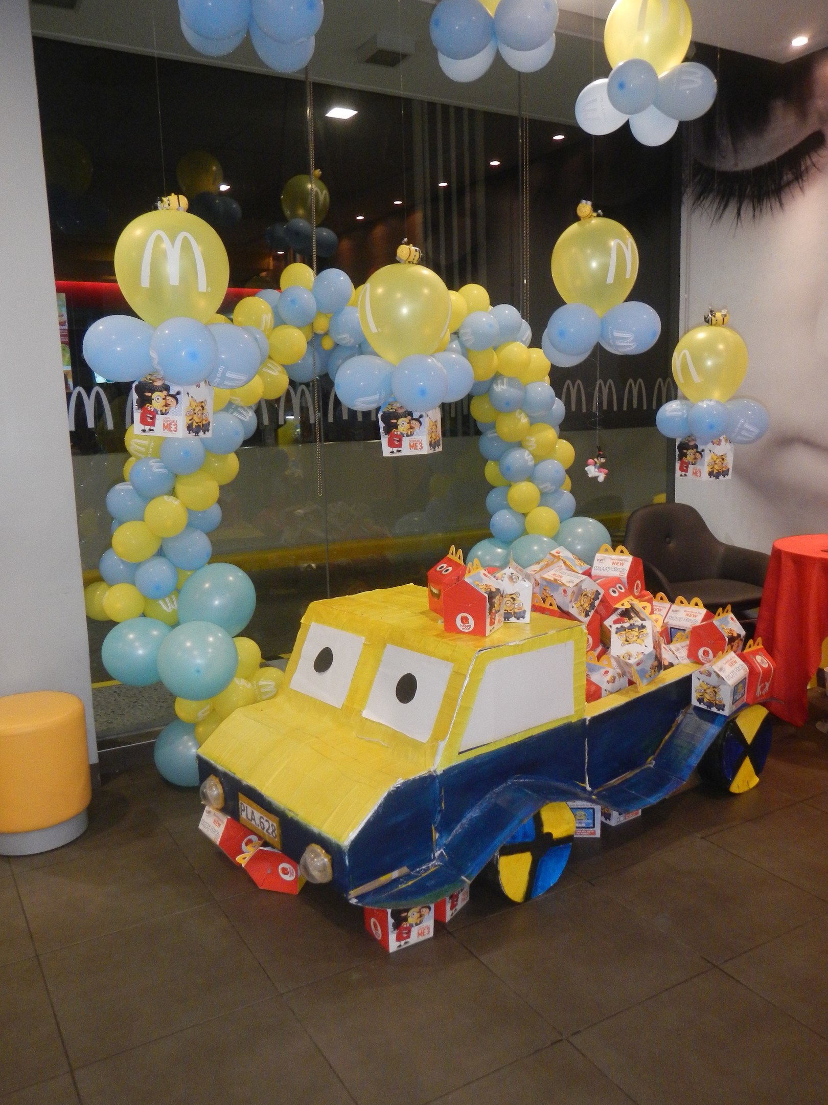 Eine McDonald's-Geburtstagsfeier mit einem gelben und blauen Lastwagen, der mit Ballons und anderen Dekoartikeln geschmückt ist, einem rot eingedeckten Tisch mit Gegenständen, Hockern und einer Glaswand im Hintergrund sowie einer Decke mit festlichen Lichtern.