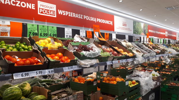 Ein Geschäftsraum mit verschiedenen Obst- und Gemüsesorten in Kartonschachteln mit Preisschildern, Informationsschilder an den Wänden und Deckenbeleuchtung.