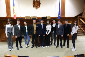 Gruppe von strahlenden Menschen, die vor einem Gerichtssaal mit Monitoren und Mikrofonen im Vordergrund und einer hölzernen Plattform mit Fahnen und Stühlen im Hintergrund posieren.