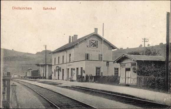 Schwarz-weiß-Foto eines Bahnhofs in Dielkirchen, Deutschland, mit einem Zug auf den Gleisen, umgeben von Häusern, Pfählen, Bäumen, Pflanzen und Menschen, Hügel im Hintergrund und einem klaren Himmel.