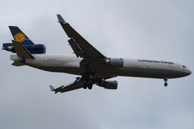 Eine Lufthansa Cargo Boeing 777-300ER im Flug, mit ausgefahrenen Flügeln, aktivierten Triebwerken und sichtbarem Text auf dem Rumpf gegen einen bewölkten Himmel.