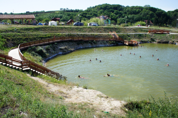 Menschen schwimmen in einem Gewässer umgeben von Grün, mit einer Brücke, Treppen, Schuppen, Fahrzeugen, Pfählen und einem klaren blauen Himmel im Hintergrund.
