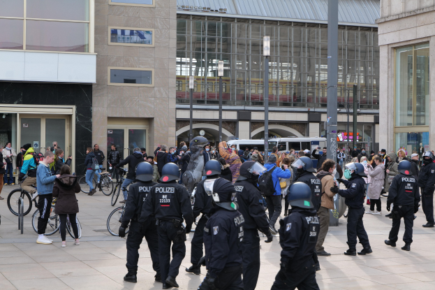 Eine Gruppe von Polizisten in Helmen steht vor einer Menge, die Fahrräder hält, mit Glasfrontgebäuden, Laternen und Fahrzeugen im Hintergrund.